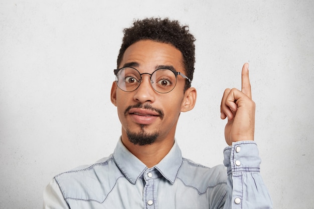 Photo gratuite jeune étudiant intelligent masculin porte des lunettes et une chemise en jean, lève l'index comme idée