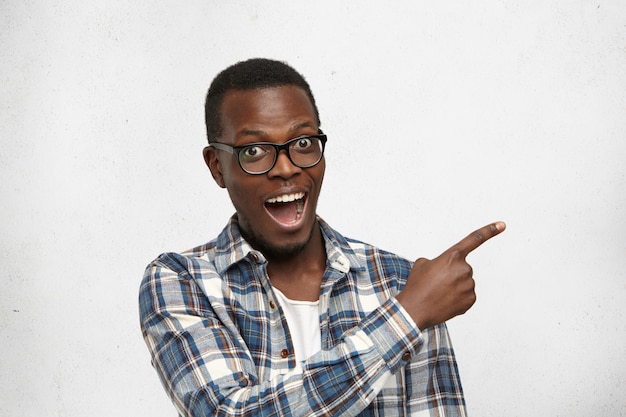 Jeune étudiant à la peau sombre excité et fasciné, portant des lunettes élégantes et une chemise à carreaux indiquant quelque chose d'étonnant