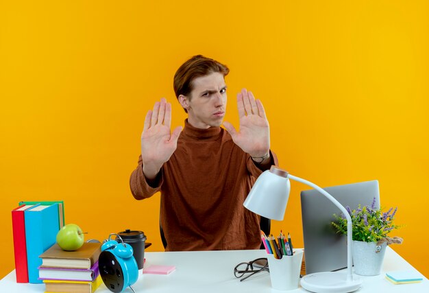 Photo gratuite jeune étudiant strict garçon assis au bureau avec des outils scolaires montrant le geste d'arrêt isolé sur un mur jaune