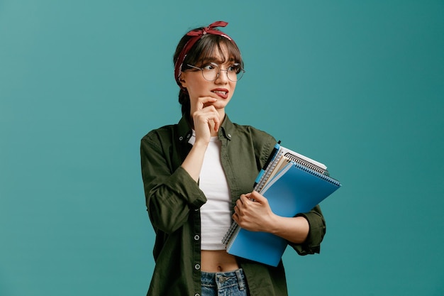 Jeune étudiante anxieuse portant des lunettes bandana tenant de grands blocs-notes avec un stylo regardant le côté touchant le menton mordant la lèvre isolé sur fond bleu