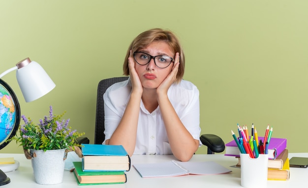 Jeune étudiante blonde confuse portant des lunettes assise au bureau avec des outils scolaires gardant les mains sur le visage en regardant la caméra isolée sur un mur vert olive