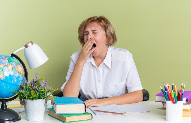Jeune étudiante blonde fatiguée assise au bureau avec des outils scolaires bâillant les yeux fermés isolé sur un mur vert olive