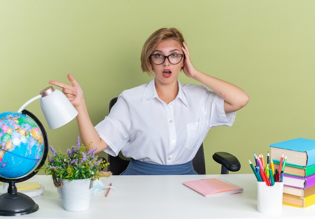 Jeune étudiante blonde inquiète portant des lunettes assise au bureau avec des outils scolaires en gardant la main sur la tête en regardant la caméra pointant sur le côté isolé sur un mur vert olive