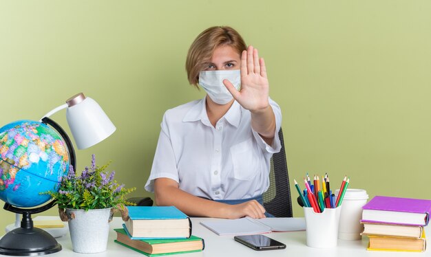 Jeune étudiante blonde sérieuse portant un masque de protection assis au bureau avec des outils scolaires regardant la caméra faisant un geste d'arrêt isolé sur un mur vert olive