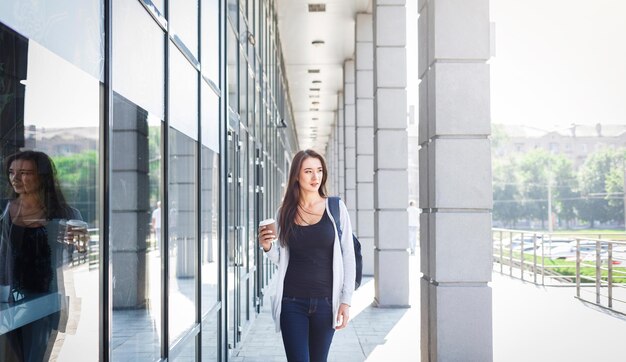 Jeune étudiante brune motivée avec sac à dos sur le chemin de l'université. boire du café avant le cours. concept de mode de vie et d'éducation