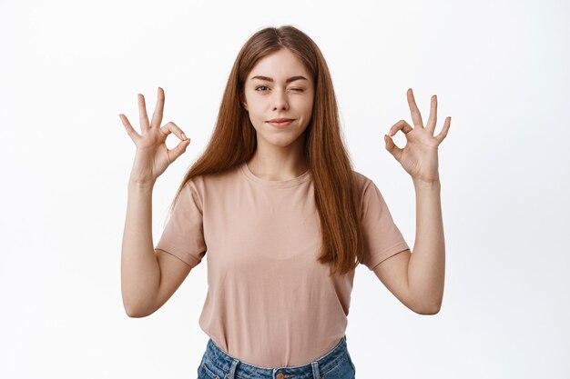 Jeune étudiante confiante faisant un clin d'œil, souriant et montrant des signes corrects, dites oui, recommandez un bon produit, approuvez ou félicitez votre choix, debout déterminé sur un mur blanc