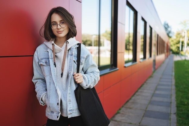 Jeune étudiante élégante queer girl appuyée sur le mur tenant un sac fourre-tout souriant regardant la caméra marchant à l'extérieur après que les cours aient terminé le programme d'éducation ou le concept de recrutement d'employés