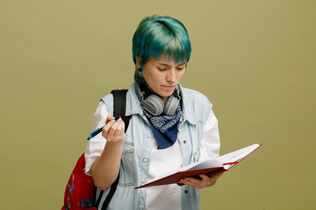 Jeune étudiante fronçant les sourcils portant des écouteurs et un bandana sur le cou et le sac à dos tenant un stylo et un carnet de notes ouvert regardant un carnet de notes isolé sur fond vert olive