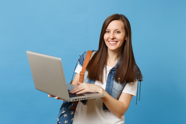 Jeune étudiante heureuse séduisante en vêtements en denim avec sac à dos tenant et utilisant un ordinateur portable isolé sur fond bleu. l'éducation au collège. copiez l'espace pour la publicité.
