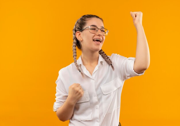 Jeune étudiante à lunettes avec des nattes en chemise blanche serrant les poings heureux et excité se réjouissant de son succès debout sur fond orange