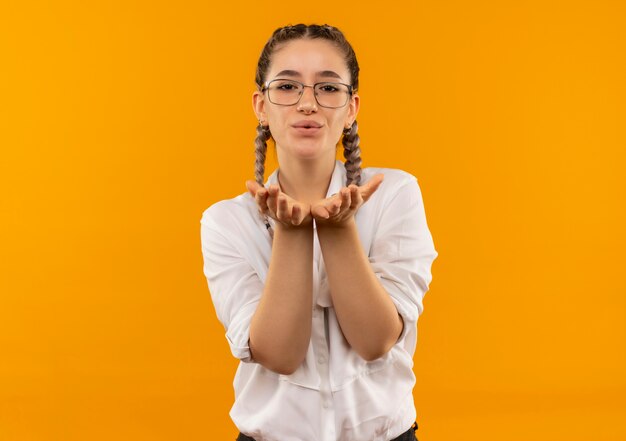 Jeune étudiante à lunettes avec des nattes en chemise blanche soufflant un baiser main dans la main devant elle debout sur un mur orange