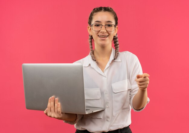 Jeune étudiante à lunettes avec des nattes en chemise blanche tenant un ordinateur portable pointant avec le doigt vers l'avant souriant joyeusement debout sur le mur rose