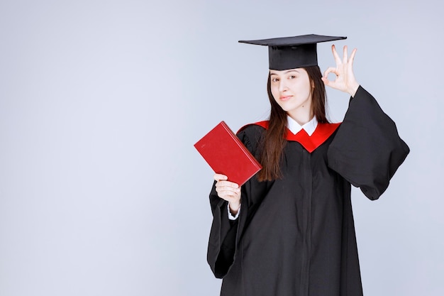 Jeune étudiante en robe académique montrant un livre rouge à la caméra. photo de haute qualité