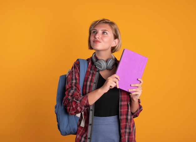 Une jeune étudiante slave impressionnée avec des écouteurs portant un sac à dos tient un livre et un cahier regardant de côté