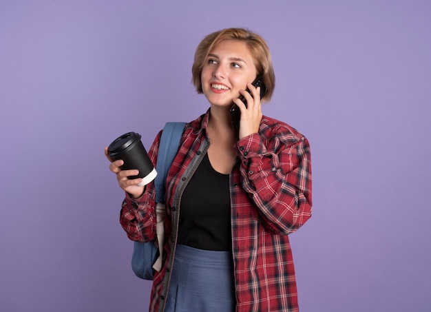 Une jeune étudiante slave souriante portant un sac à dos tient des discussions sur une tasse de papier au téléphone en regardant de côté