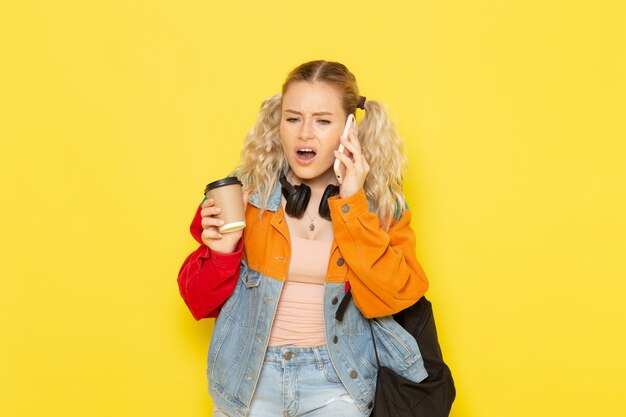 Jeune étudiante en vêtements modernes parlant au téléphone tenant du café sur jaune