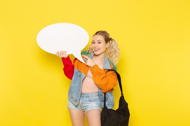 Jeune étudiante en vêtements modernes tenant un énorme panneau blanc avec sourire sur jaune