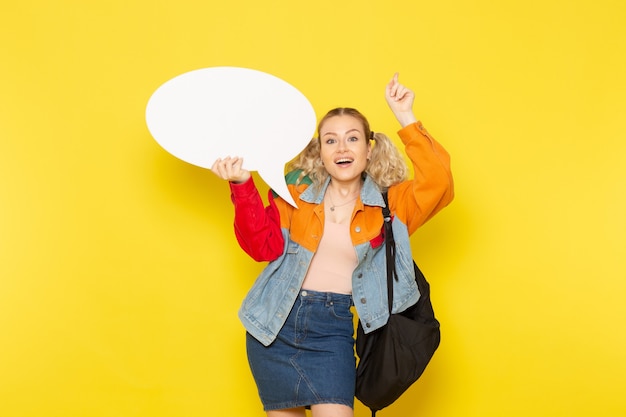 Jeune étudiante en vêtements modernes tenant un énorme panneau blanc avec sourire sur jaune
