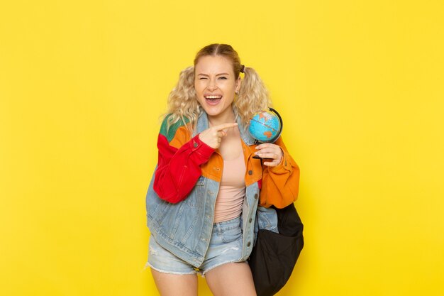 Jeune étudiante en vêtements modernes tenant petit globe souriant sur jaune