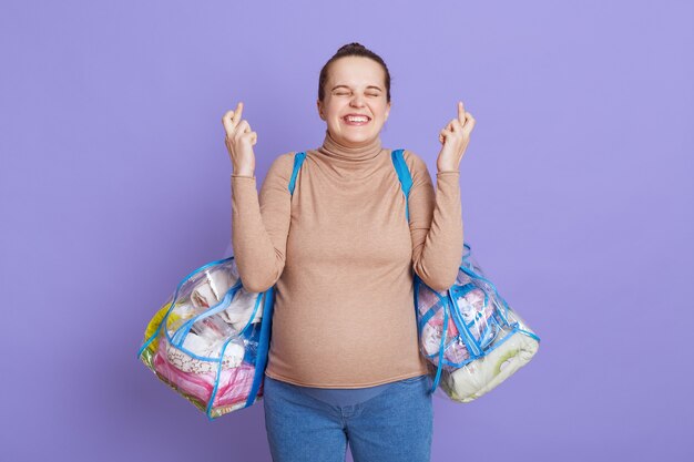 Jeune européenne belle fille enceinte avec chignon attend bébé, se tient isolé sur un mur lilas, faisant des gestes de doigt croisé, souriant, avec une expression d'espoir, tenant des trucs pour la maison de maternité.