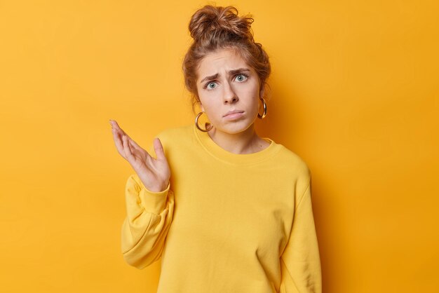 Une jeune européenne confuse et douteuse aux cheveux peignés lève la paume et a l'air perplexe réagit à quelque chose d'inattendu porte des poses de pull décontractées sur fond jaune vif Alors que faire maintenant