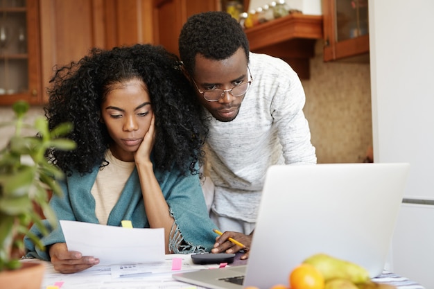 Jeune famille africaine de payer les factures de services publics en ligne à l'aide d'un ordinateur portable. Malheureuse femme assise à table analyse feuille de papier dans sa main, calcul des dépenses domestiques avec son mari