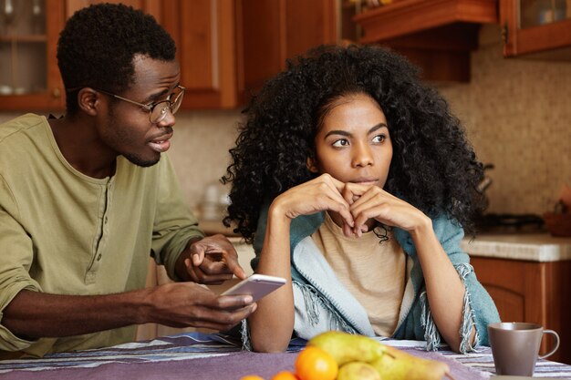 Jeune famille afro-américaine qui se bat dans la cuisine à cause d'une liaison. Homme à lunettes tenant un téléphone portable, pointant le doigt sur l'écran, essayant de s'expliquer pour les messages d'amour d'une femme inconnue