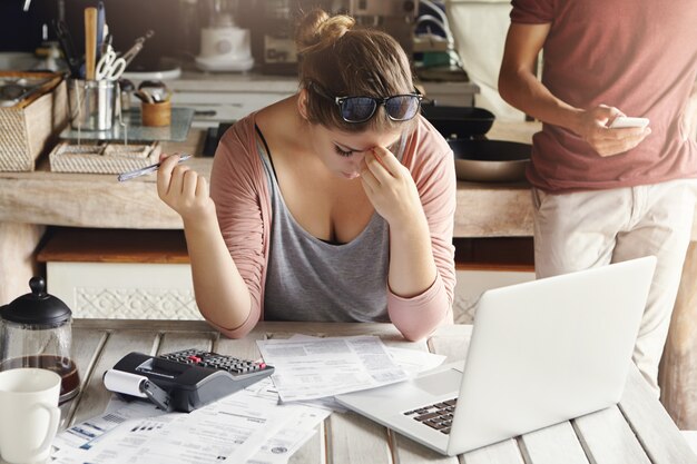 Jeune famille confrontée à des problèmes d'endettement. Femme stressée tenant sa tête dans la frustration, faire des comptes bancaires et effectuer les calculs nécessaires à l'aide d'un ordinateur portable et d'une calculatrice