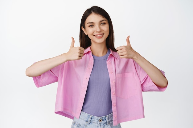 Photo gratuite jeune femme de 25 ans montre le pouce vers le haut recommande que le magasin donne son approbation en souriant et en disant oui debout sur fond blanc