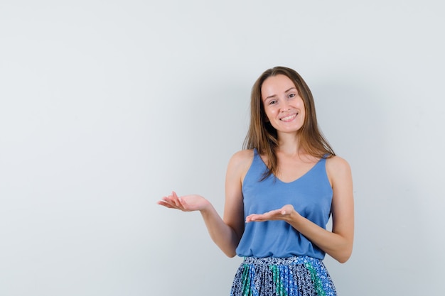 Jeune femme accueillant ou montrant quelque chose en maillot, jupe et à la recherche de bonne humeur. vue de face.