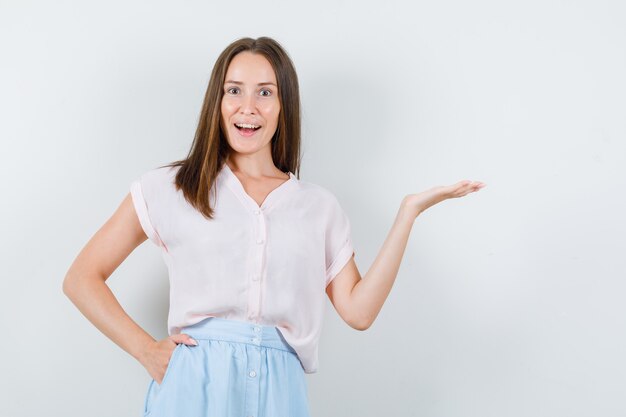 Jeune femme accueillant ou montrant quelque chose en t-shirt, jupe et à l'optimiste. vue de face.