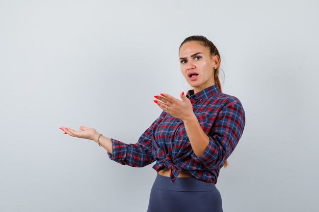 Jeune femme accueillant quelque chose en chemise à carreaux, pantalon et à la perplexité. vue de face.