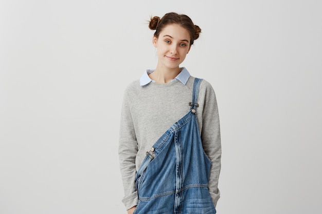 Jeune femme adorable à la recherche de combinaison denim décontractée. Dame brune affable aux cheveux en double chignon profitant du temps passé à la maison le week-end. Concept de confort