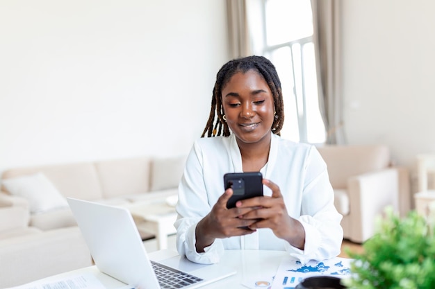 Jeune femme d'affaires africaine souriante utilisant un smartphone près d'un ordinateur au bureau