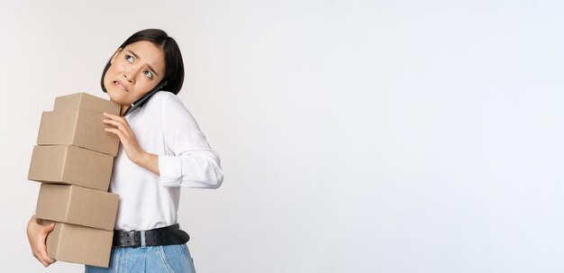 Une jeune femme d'affaires asiatique répond à un appel téléphonique en parlant sur son mobile tout en transportant une pile de boîtes avec des commandes debout sur fond blanc