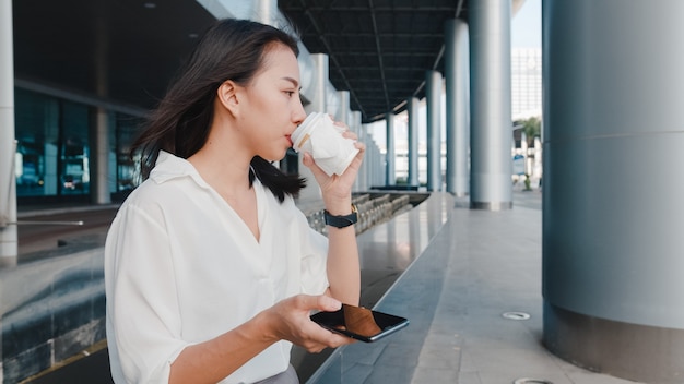 Jeune femme d'affaires asiatique réussie dans des vêtements de bureau de mode tenant une tasse de papier jetable de boisson chaude et utilisant un téléphone intelligent tout en se tenant à l'extérieur dans une ville moderne urbaine