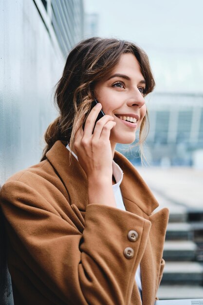 Jeune femme d'affaires assez élégante en manteau parlant joyeusement sur un téléphone portable dans la rue de la ville