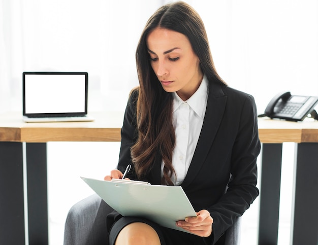 Jeune femme d&#39;affaires assis devant le bureau écrit sur le presse-papiers avec stylo