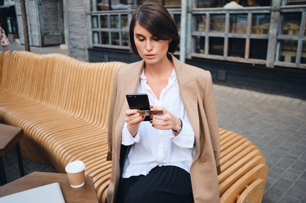 Jeune femme d'affaires élégante et confiante avec carte de crédit à l'aide d'un téléphone portable pendant la pause-café au café dans la rue