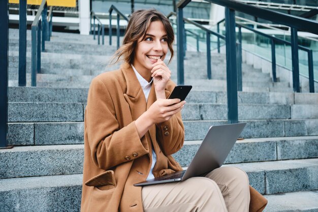 Jeune femme d'affaires excitée positive en manteau utilisant rêveusement un téléphone portable tout en travaillant avec un ordinateur portable dans les escaliers en plein air