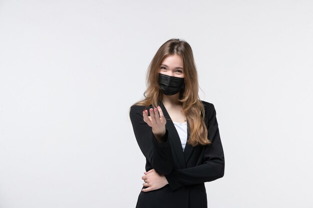 Jeune femme d'affaires heureuse en costume portant un masque chirurgical et pointant quelqu'un sur un mur blanc isolé