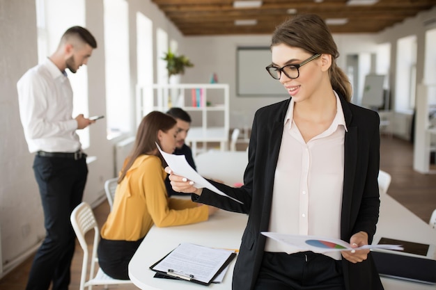 Jeune femme d'affaires à lunettes et chemise tenant des documents à la main tout en travaillant au bureau avec des collègues en arrière-plan