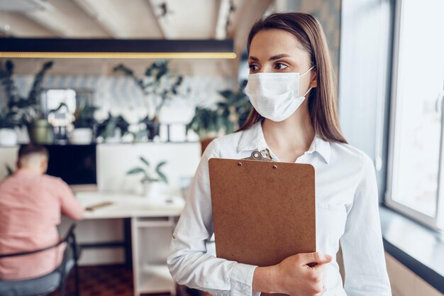 Jeune femme d'affaires en masque facial debout au bureau et tenant un presse-papiers avec des documents