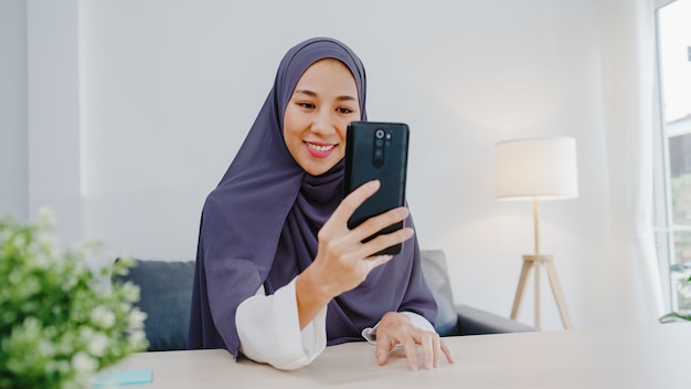 Une jeune femme d'affaires musulmane utilisant un téléphone intelligent parle à un ami par vidéoconférence lors d'une réunion en ligne tout en travaillant à distance depuis la maison dans le salon.