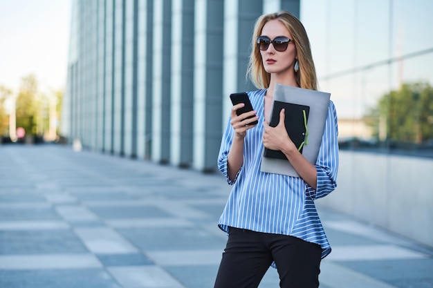 Une jeune femme d'affaires prospère attend son nouveau partenaire dans la rue avec un téléphone portable dans les mains.