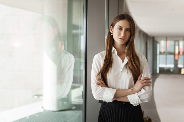 Jeune femme d'affaires prospère, debout près de la fenêtre dans le couloir du bureau, souriant et regardant la caméra.