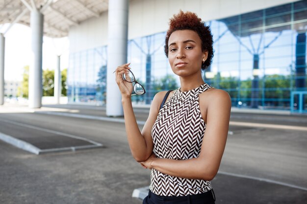 Jeune femme d'affaires prospère tenant des lunettes, debout près du centre d'affaires