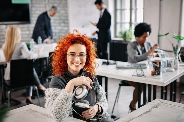 Jeune femme d'affaires rousse heureuse tenant des écouteurs et regardant la caméra alors qu'elle était assise à son bureau au bureau Ses collègues travaillent en arrière-plan