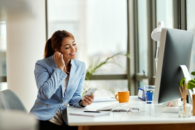 Jeune femme d'affaires se détendre tout en utilisant un téléphone portable et en écoutant de la musique sur des écouteurs au bureau