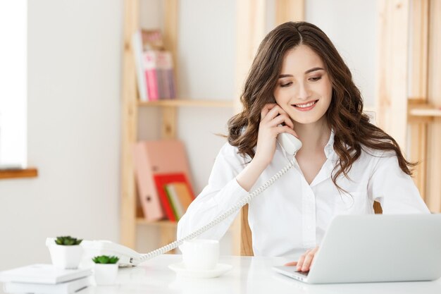 Jeune femme d'affaires ou secrétaire assis au bureau et travaillant souriant et regardant la caméra